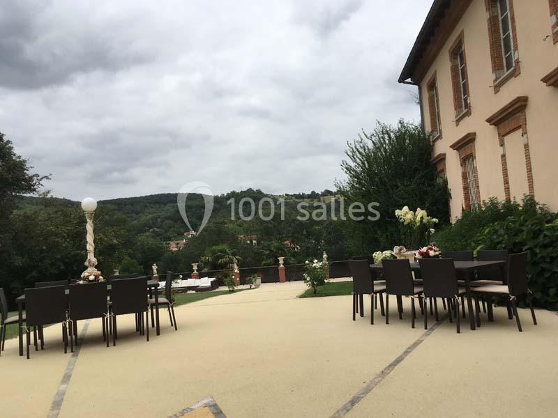 Terrasse extérieure avec tables et chaises, vue sur un jardin verdoyant et des collines sous un ciel nuageux.
