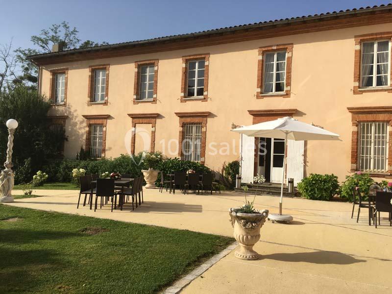 Façade d'un bâtiment ancien avec terrasse aménagée, tables, chaises, parasol et végétation environnante.