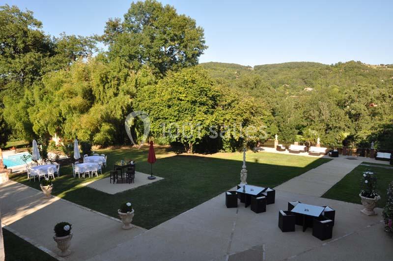 Jardin avec pelouse, tables et chaises disposées, entouré d'arbres et collines en arrière-plan.