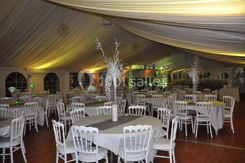 Salle de réception sous chapiteau avec tables rondes dressées, chaises blanches et décorations lumineuses.