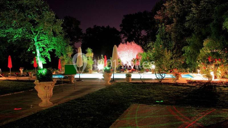 Jardin éclairé de nuit avec piscine entourée de végétation, parasols et chaises longues.