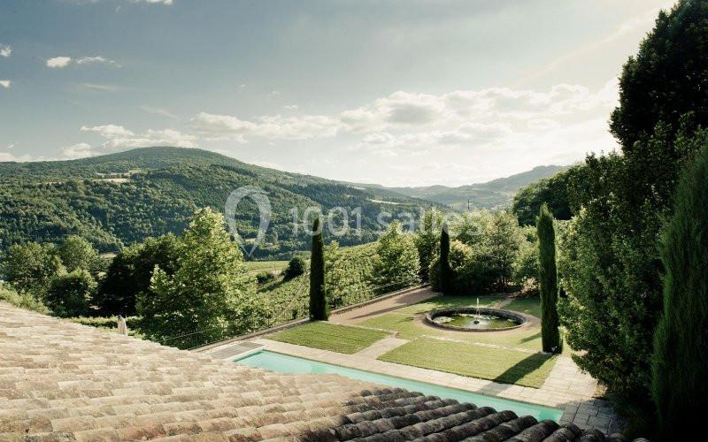 Vue sur un paysage vallonné avec des arbres, une piscine, une fontaine et un toit en tuiles au premier plan.