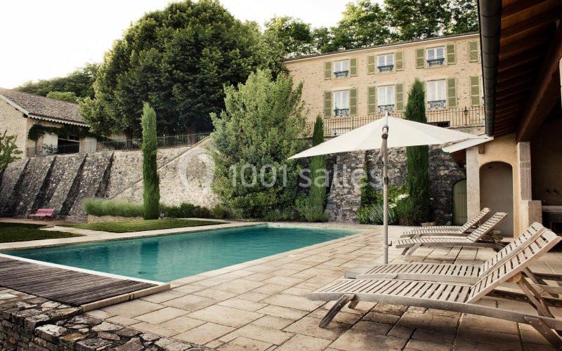 Piscine extérieure entourée de chaises longues en bois, située devant une maison en pierre avec des arbres en arrière-plan.
