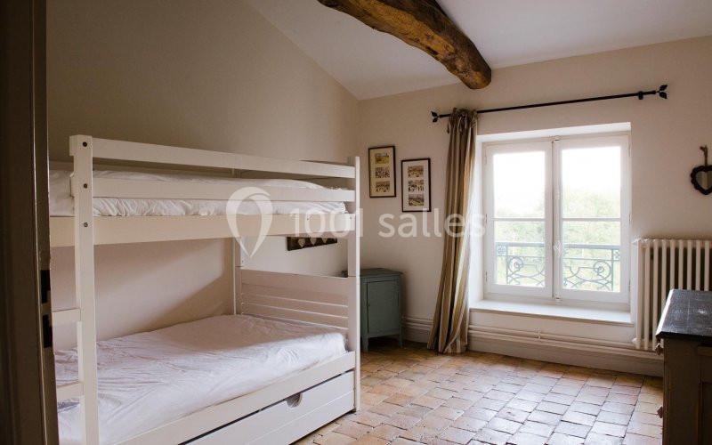 Chambre lumineuse avec lits superposés blancs, fenêtre à double battant, sol en terre cuite et poutre apparente.