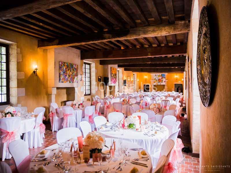 Salle de réception décorée avec des tables rondes, nappes blanches, chaises ornées de rubans roses et lumière naturelle.