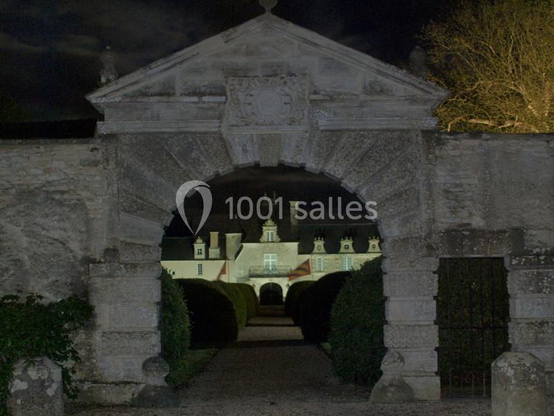 Arc en pierre encadrant une allée menant à un château éclairé de nuit, entouré de haies et d'arbres.