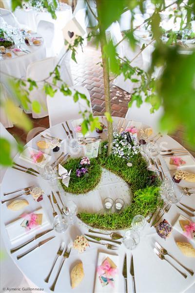 Table ronde décorée pour un événement, ornée de mousse, fleurs et un arbre central, avec vaisselle disposée autour.