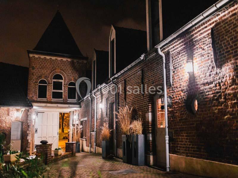 Façade en briques d'un bâtiment éclairé la nuit, avec des plantes en pots et une porte blanche.