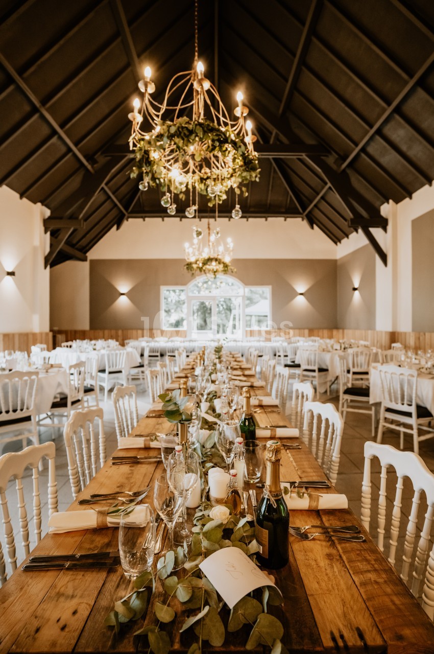 Salle de réception décorée avec des tables en bois, chandeliers suspendus et chaises blanches alignées.