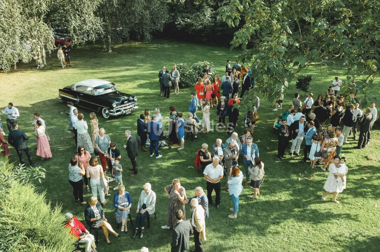 Groupe de personnes rassemblées dans un jardin verdoyant, avec une voiture ancienne noire en arrière-plan.