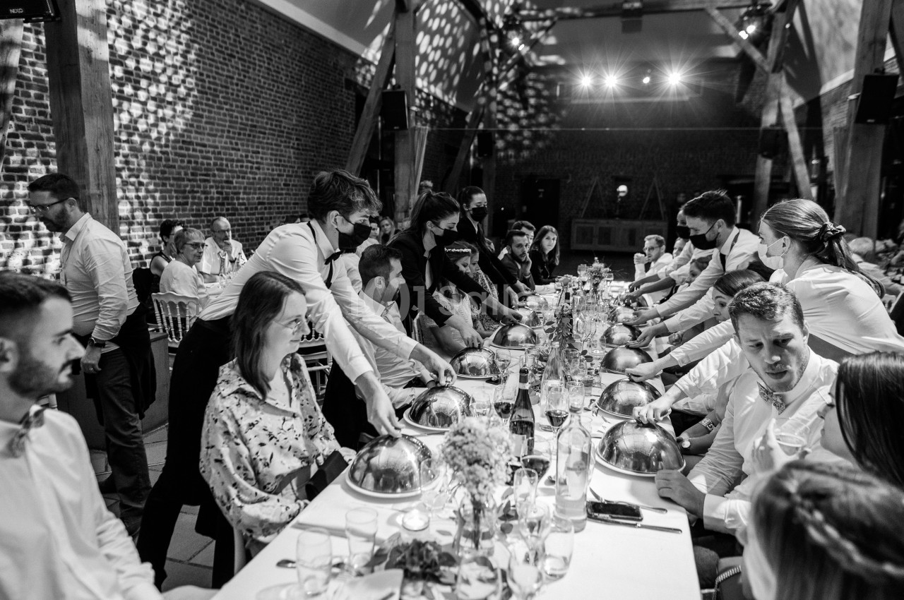 Des serveurs déposent des plats sous cloches sur une longue table lors d'un dîner dans une salle en briques.