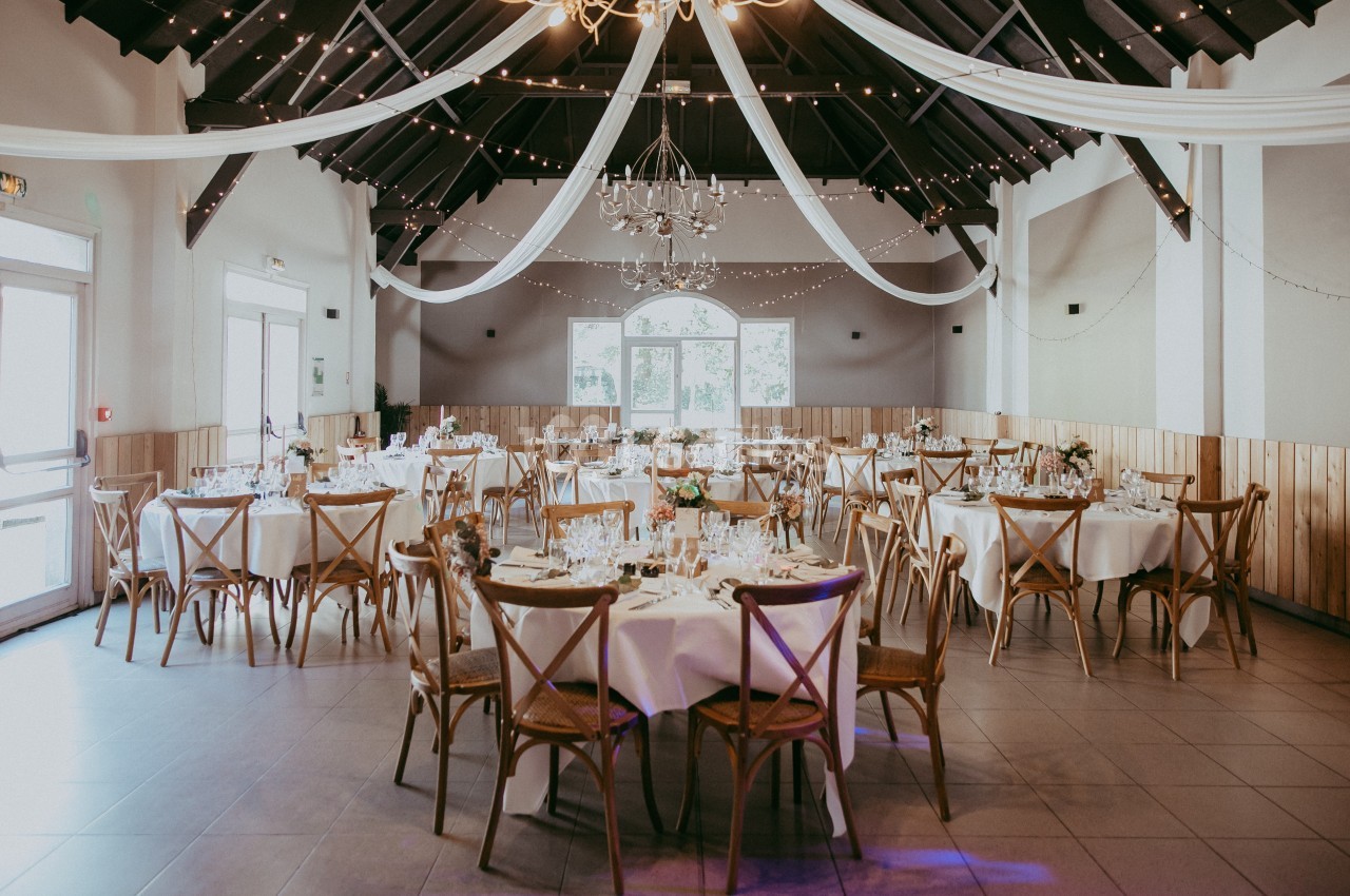 Salle de réception décorée avec des tables rondes dressées, des chaises en bois et des guirlandes lumineuses suspendues.