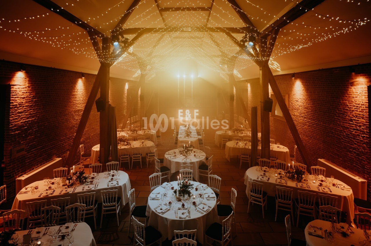 Salle de réception décorée avec des guirlandes lumineuses, tables rondes dressées et initiales éclairées en arrière-plan.