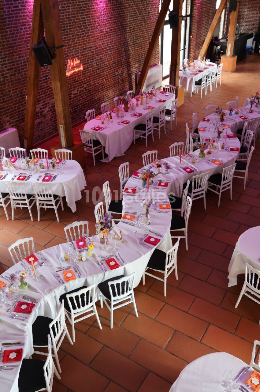 Salle de réception avec tables dressées en U, nappes blanches, chaises blanches et décorations florales colorées.