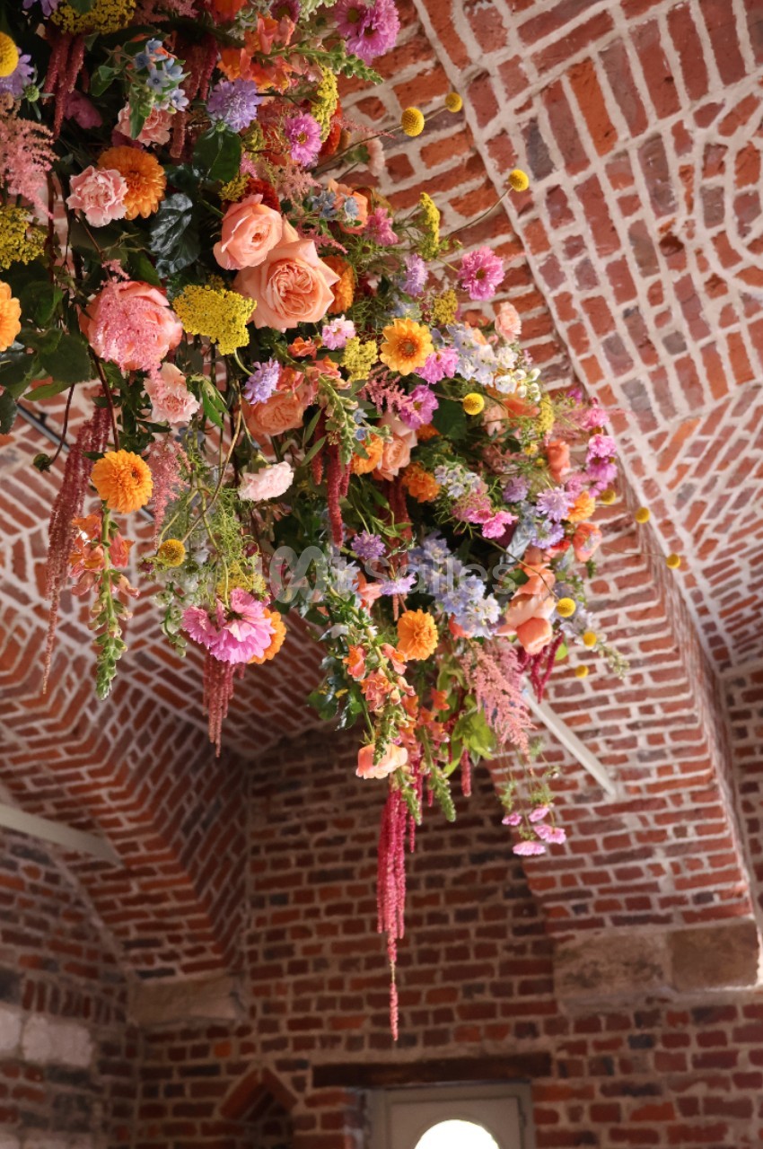 Suspension florale colorée avec des roses, des marguerites et des feuillages, accrochée sous une voûte en briques.
