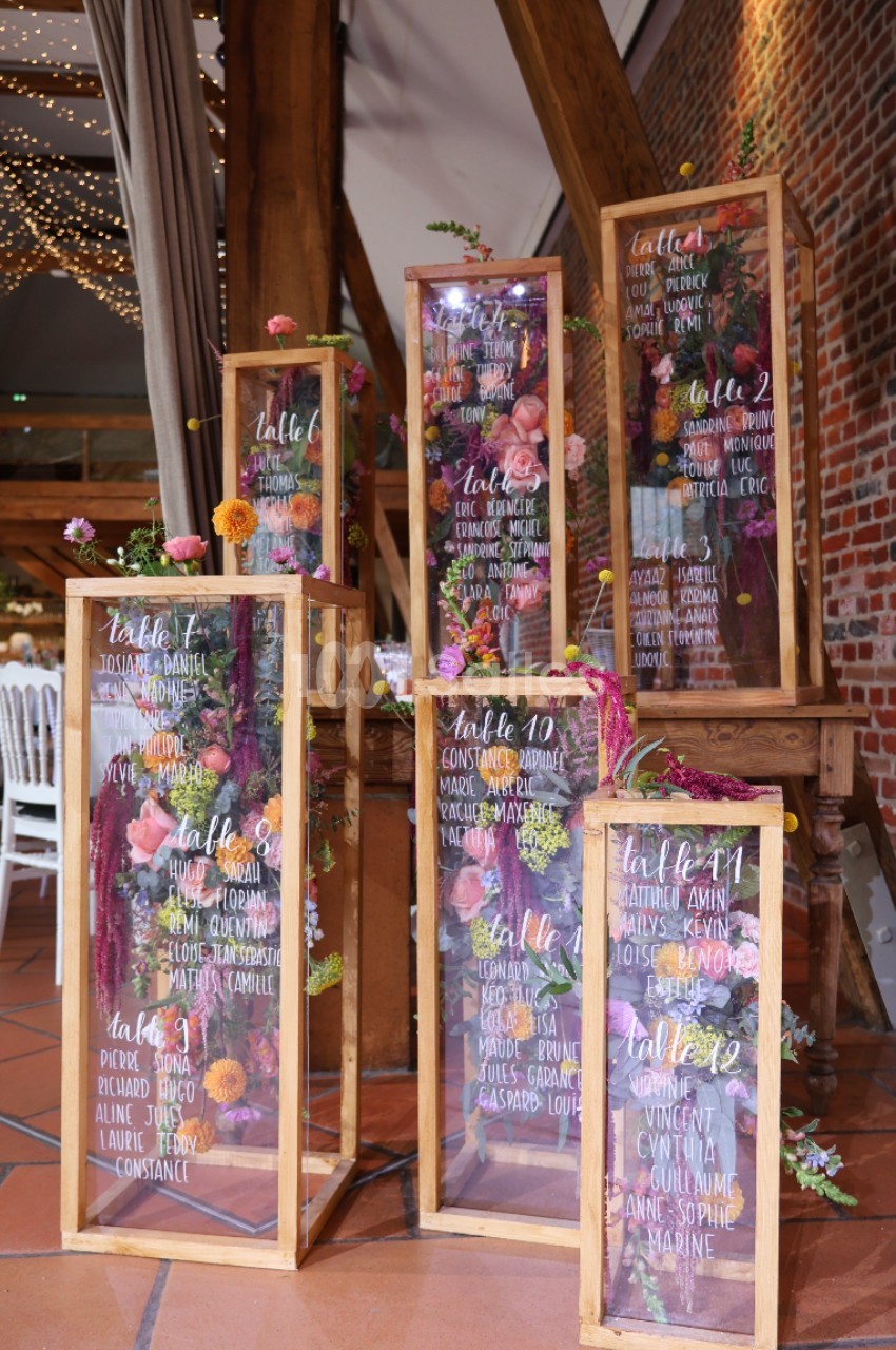 Panneaux de plan de table en verre décorés de fleurs colorées, disposés dans des cadres en bois dans une salle de réception.