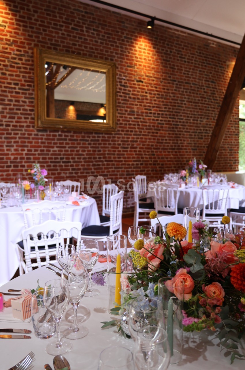 Salle de réception décorée avec des tables rondes dressées, des fleurs colorées et un mur de briques apparentes.