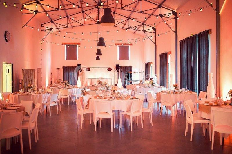Salle de réception décorée avec des guirlandes lumineuses, tables rondes dressées et chaises blanches.
