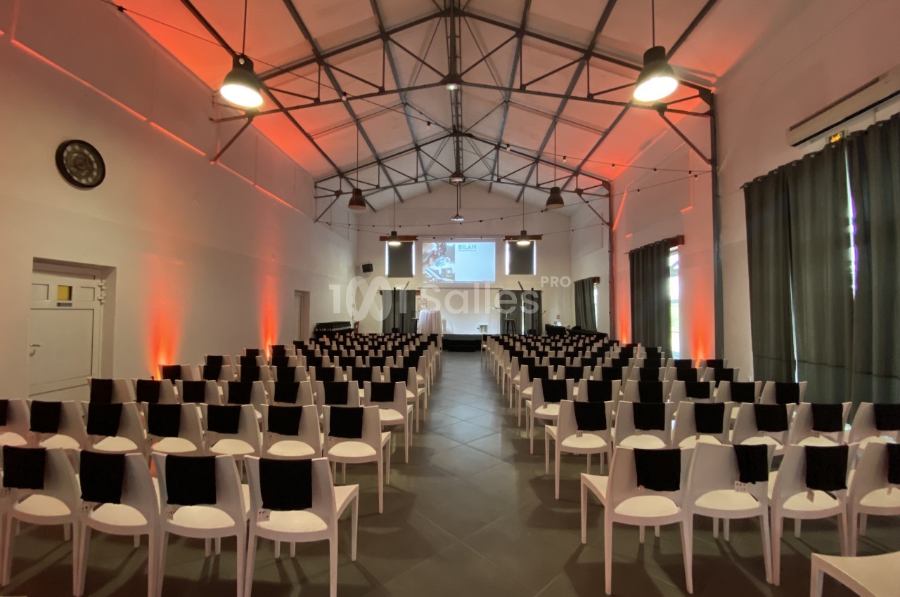 Salle de conférence avec rangées de chaises blanches, écran de projection et éclairage tamisé orange.