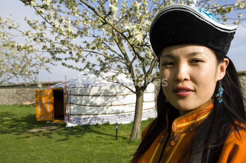 Femme en tenue traditionnelle devant une yourte blanche et un arbre en fleurs dans un espace extérieur verdoyant.