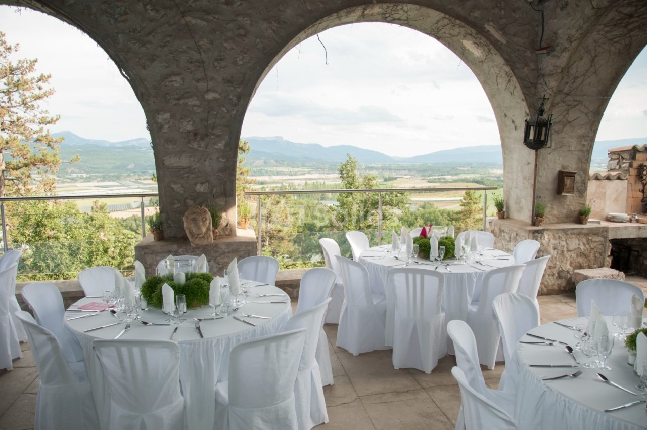 Terrasse avec tables rondes dressées, nappes blanches, vue sur une vallée verdoyante et montagnes en arrière-plan.
