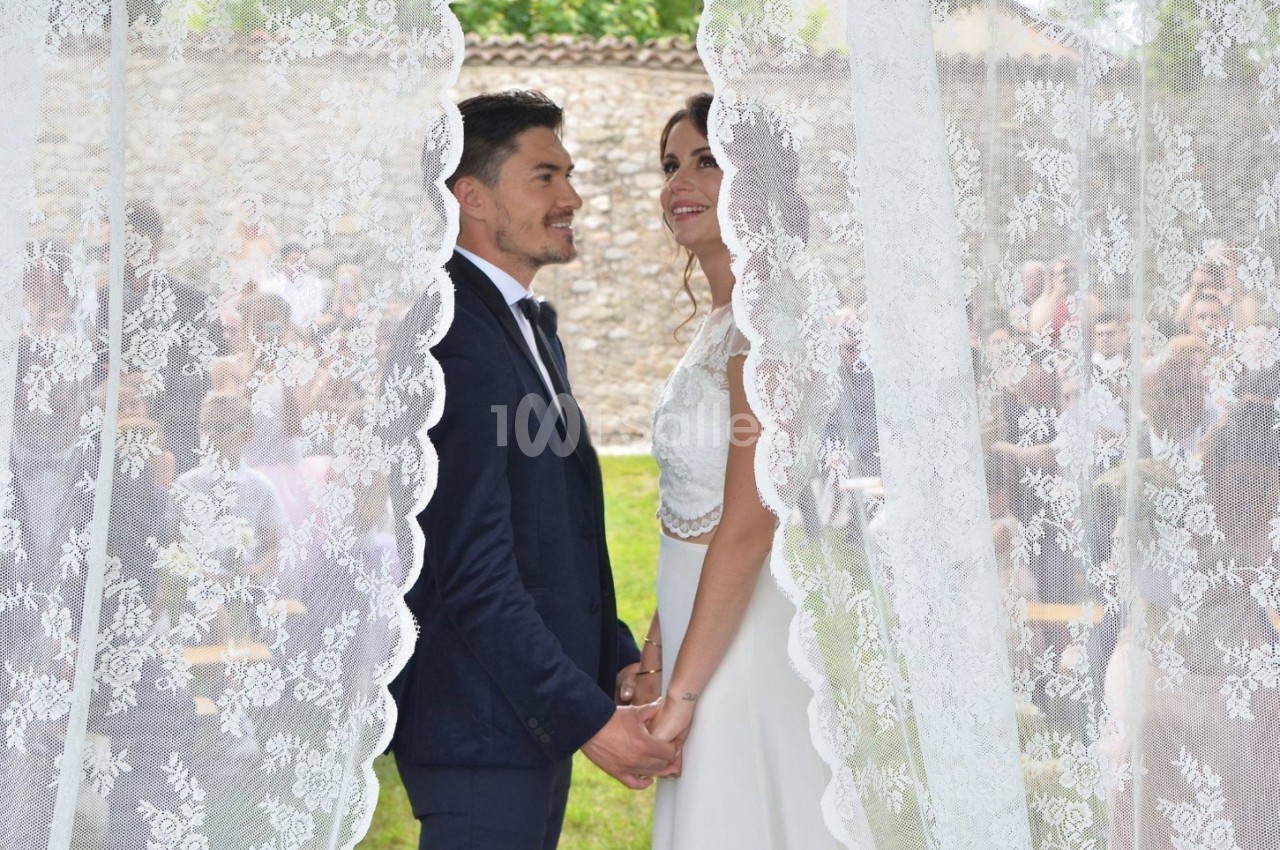 Un couple en tenue de mariage se tient la main, encadré par des rideaux en dentelle, avec des invités en arrière-plan flou.
