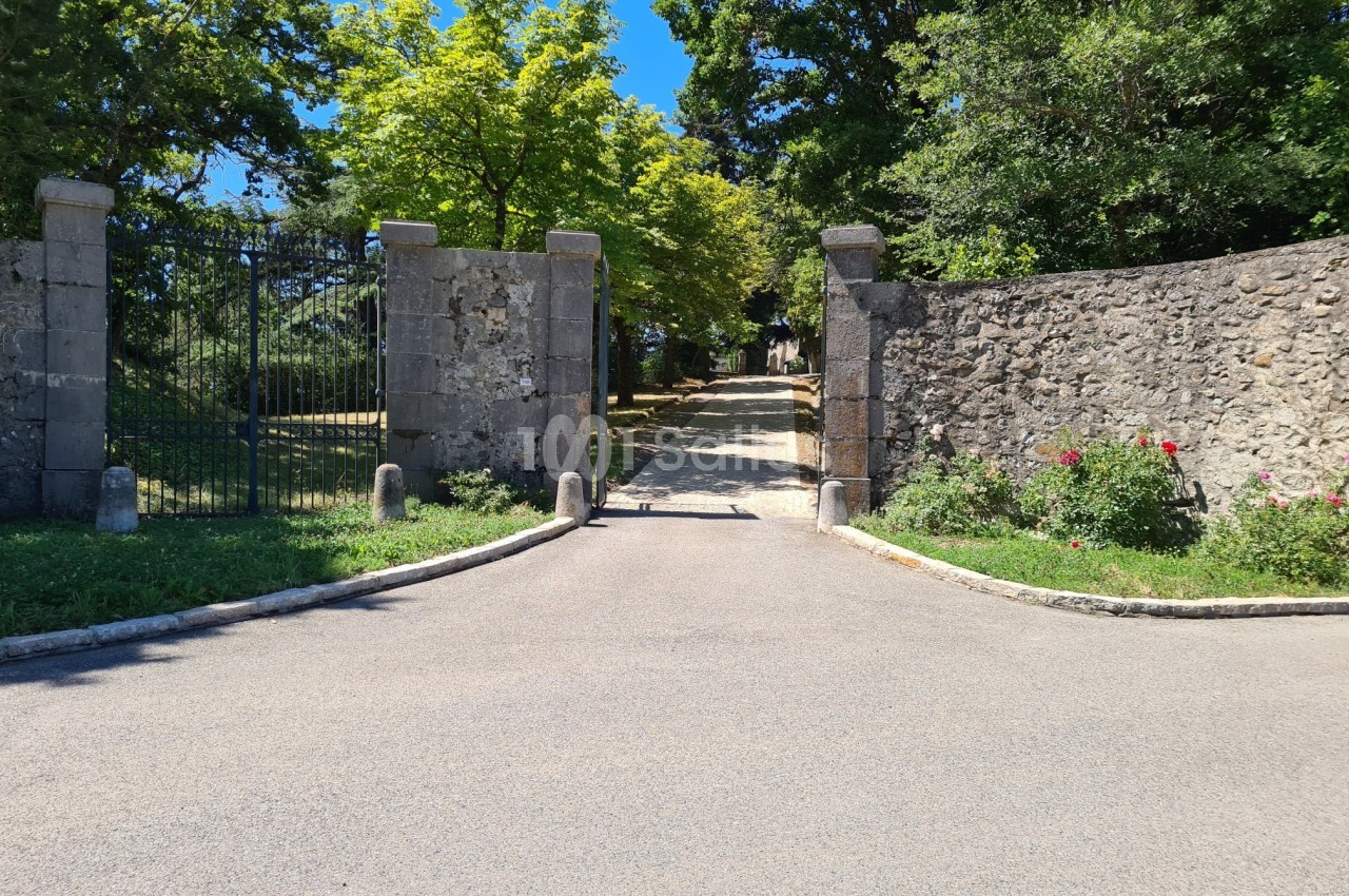 Entrée d'un chemin bordé de murs en pierre et de végétation, avec un portail ouvert sous un ciel dégagé.