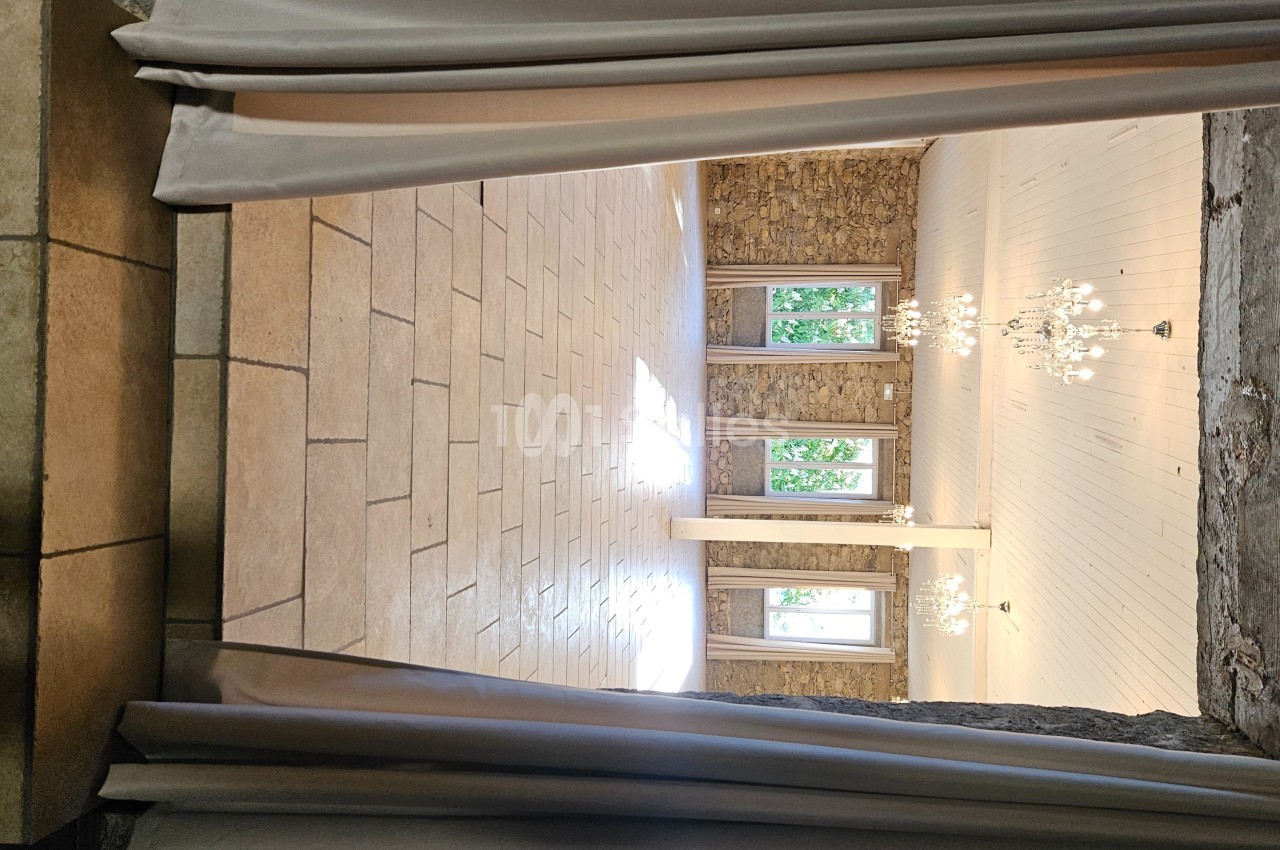 Salle lumineuse avec murs en pierre, carrelage beige, lustres suspendus et grandes fenêtres donnant sur l'extérieur.