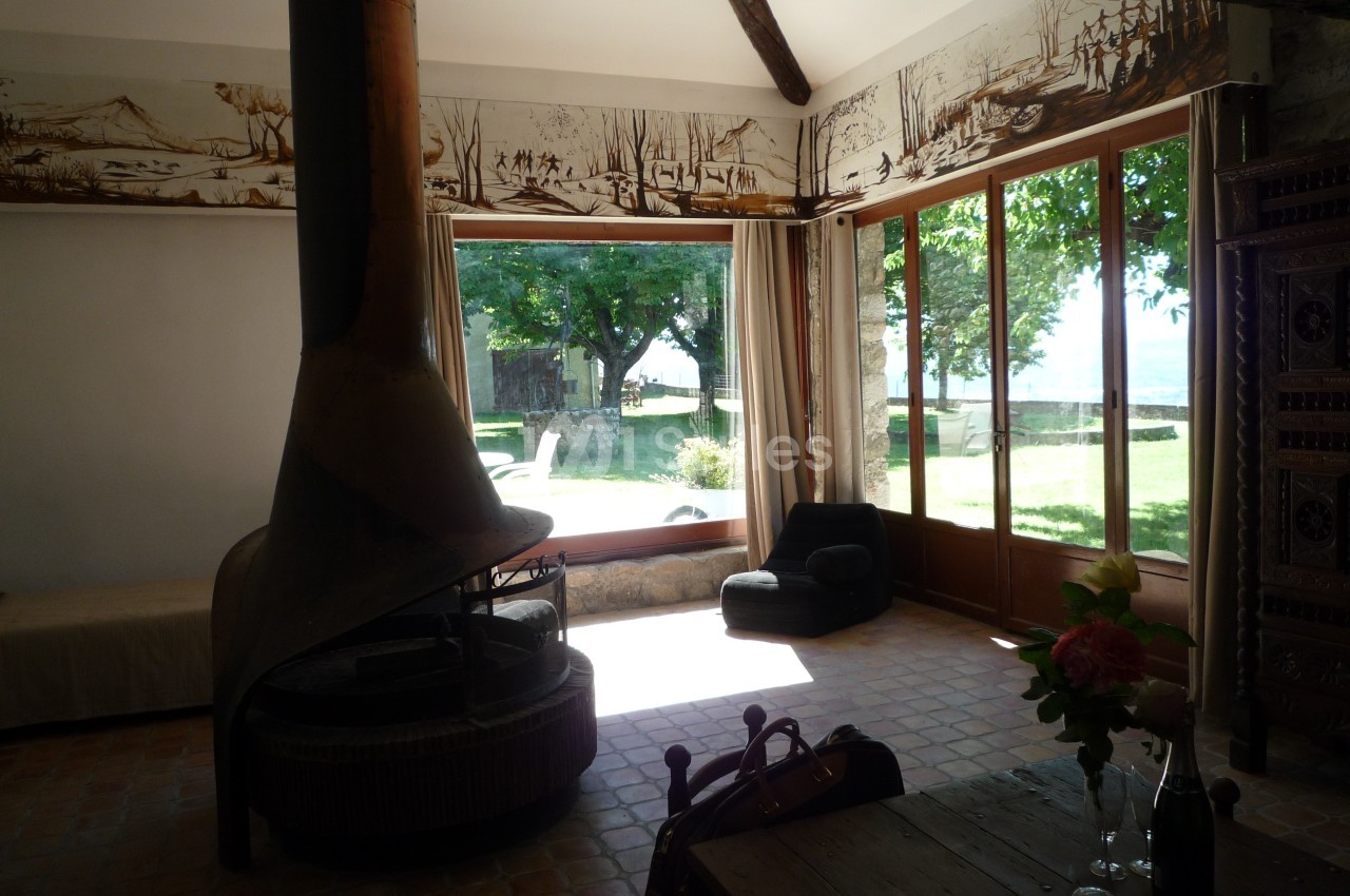 Salon lumineux avec cheminée centrale, grandes baies vitrées donnant sur un jardin arboré et mobilier rustique.