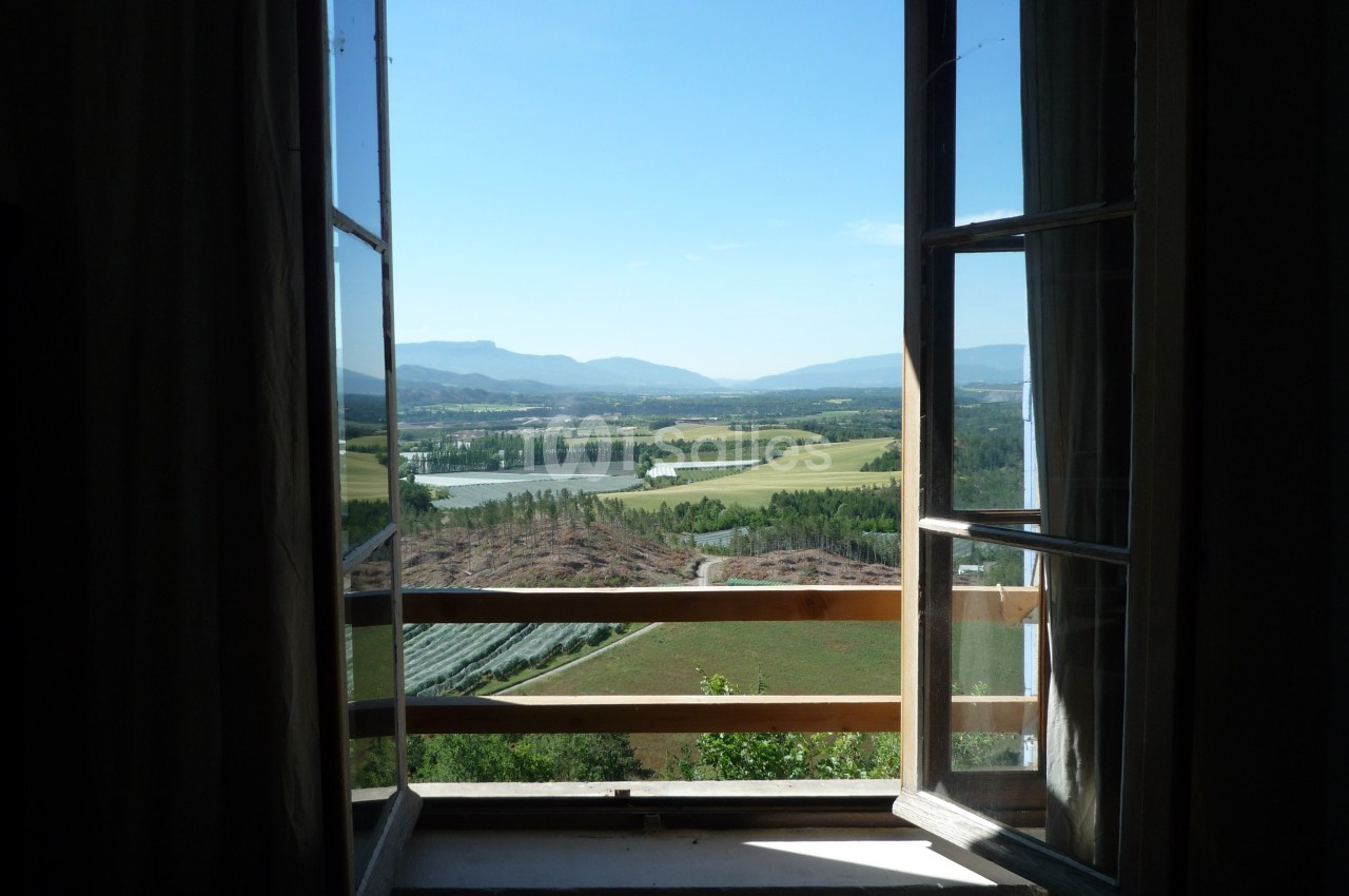 Vue depuis une fenêtre ouverte sur un paysage rural avec champs, collines et un ciel dégagé.