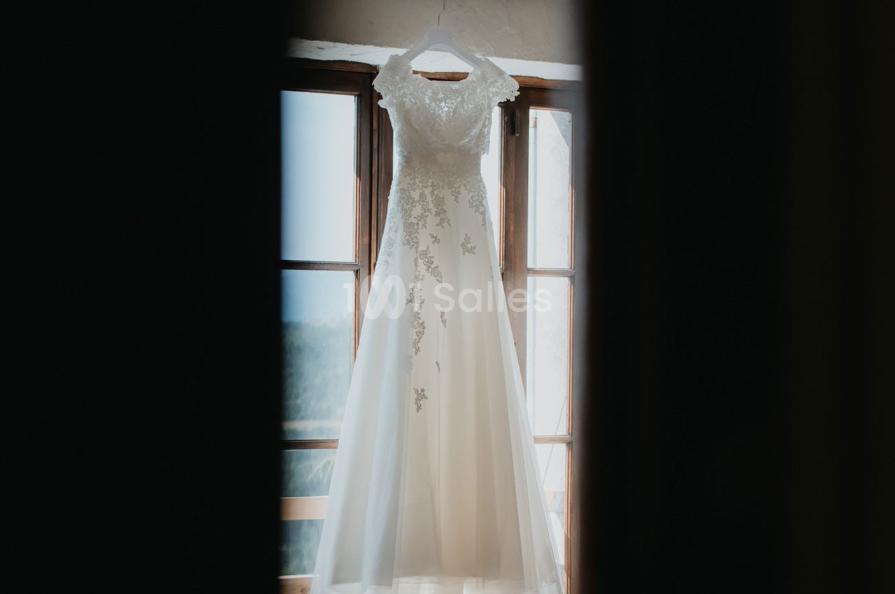 Robe de mariée blanche ornée de dentelle suspendue devant une fenêtre lumineuse.