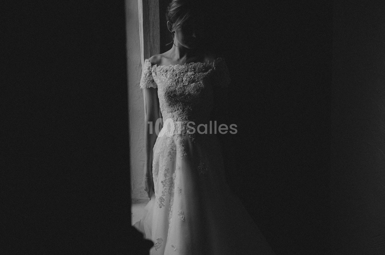 Femme en robe de mariée en dentelle, debout près d'une fenêtre dans une ambiance sombre et tamisée.