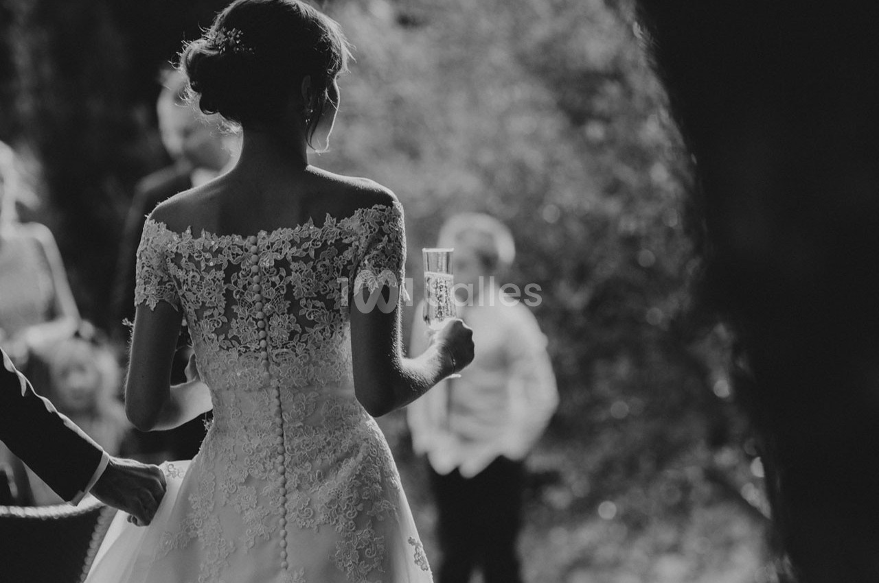Une mariée en robe blanche ornée de dentelle tient un verre, vue de dos dans un cadre extérieur flou.