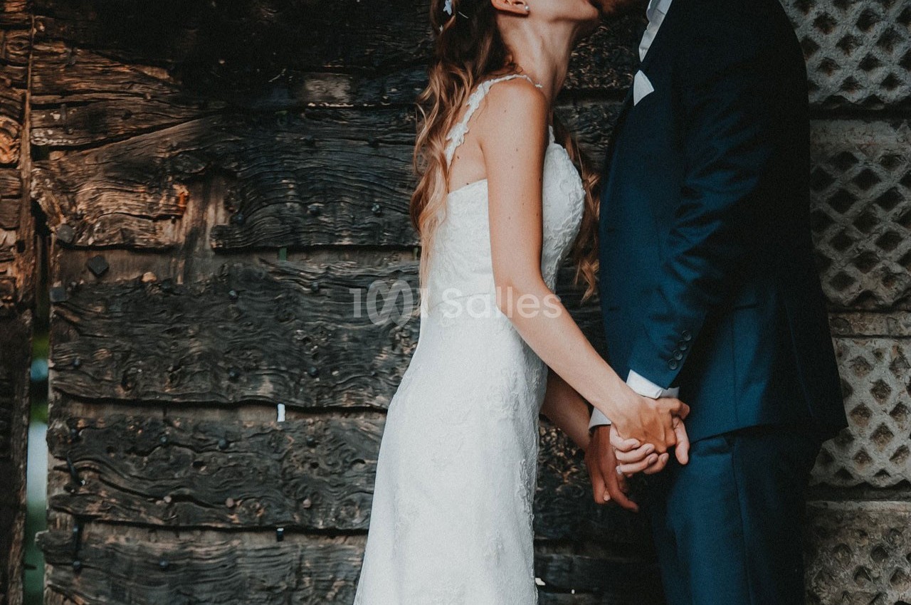 Un couple en tenue de mariage s'embrasse devant un mur en bois rustique, se tenant les mains.