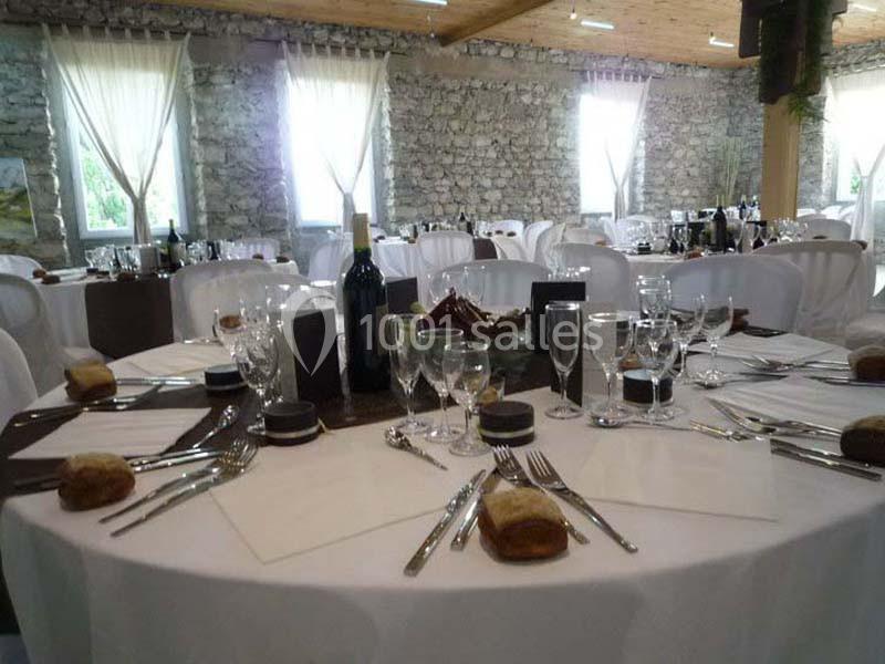Table dressée pour un repas, avec nappes blanches, couverts, verres, pain et bouteilles de vin, dans une salle en pierre.