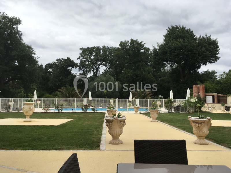 Vue d'une piscine extérieure entourée de chaises longues, parasols et végétation sous un ciel nuageux.