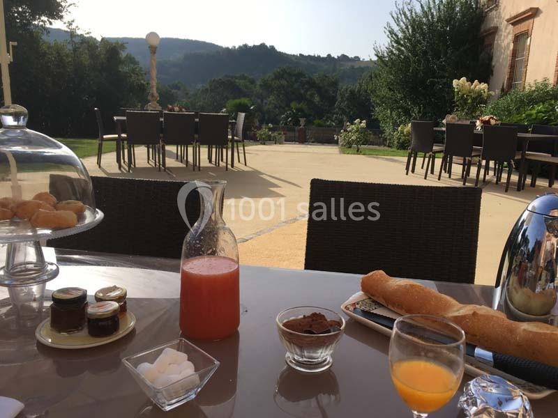 Petit-déjeuner en terrasse avec jus, confitures, viennoiseries et vue sur un jardin verdoyant et des collines.