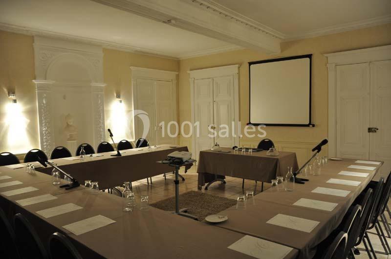 Salle de réunion éclairée avec tables disposées en U, chaises, projecteur, écran blanc et micros.