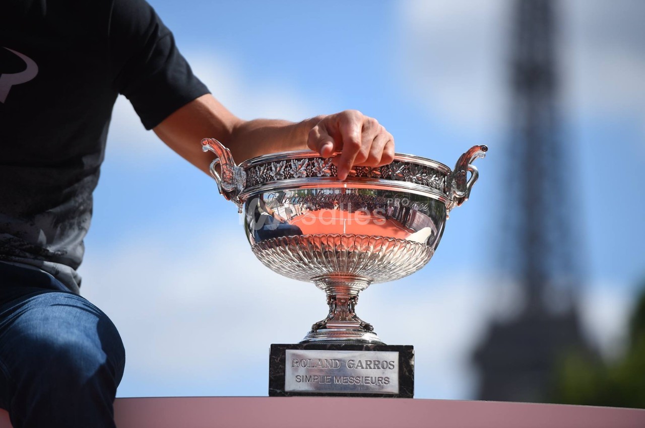 Une personne touche un trophée en argent marqué ’Roland Garros Simple Messieurs’, avec la Tour Eiffel en arrière-plan.