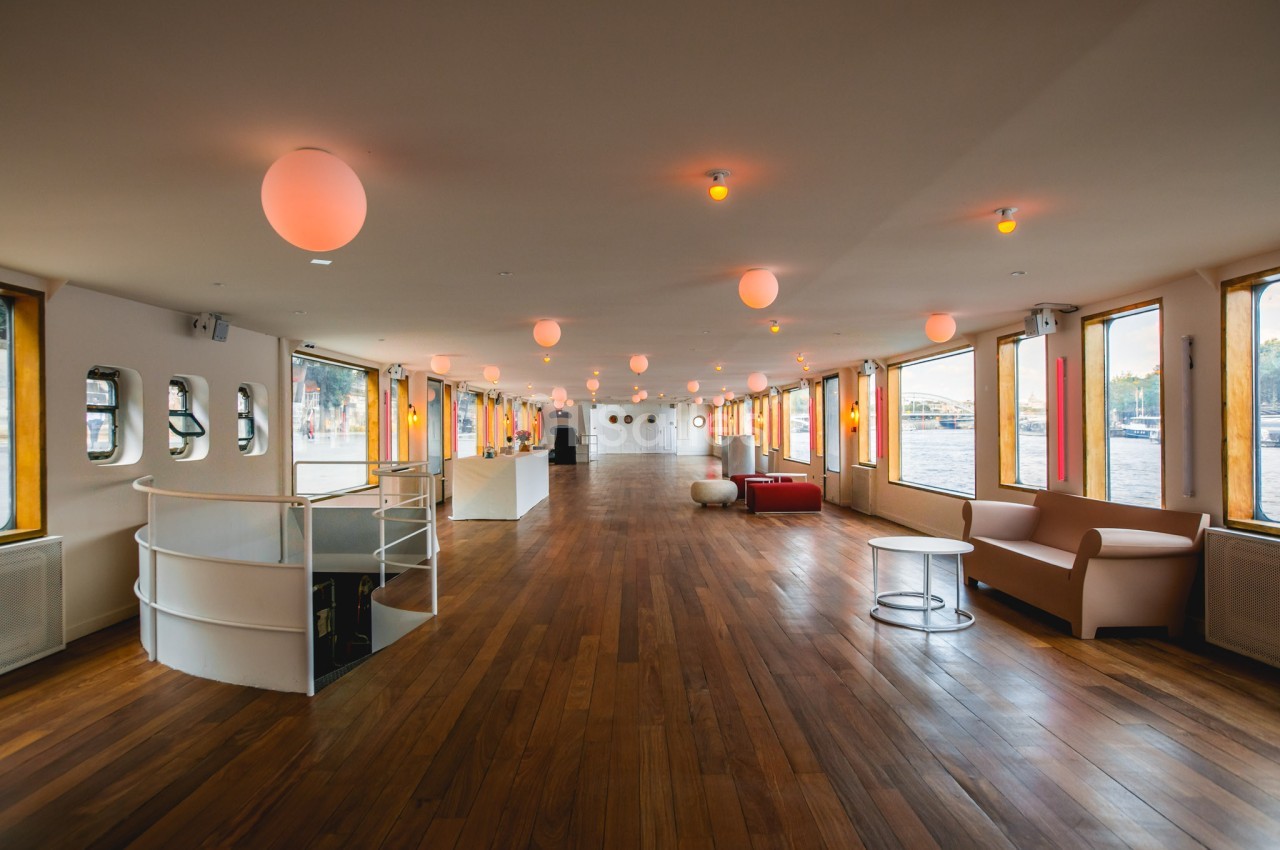 Salle spacieuse avec parquet en bois, canapés, baies vitrées et éclairage suspendu, offrant une vue sur l'extérieur.