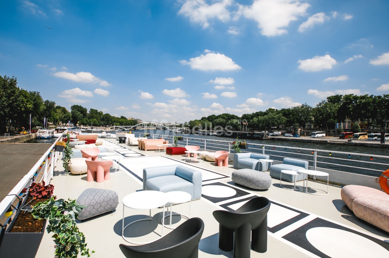 Terrasse aménagée avec des fauteuils et tables modernes, offrant une vue sur un fleuve et un pont sous un ciel dégagé.
