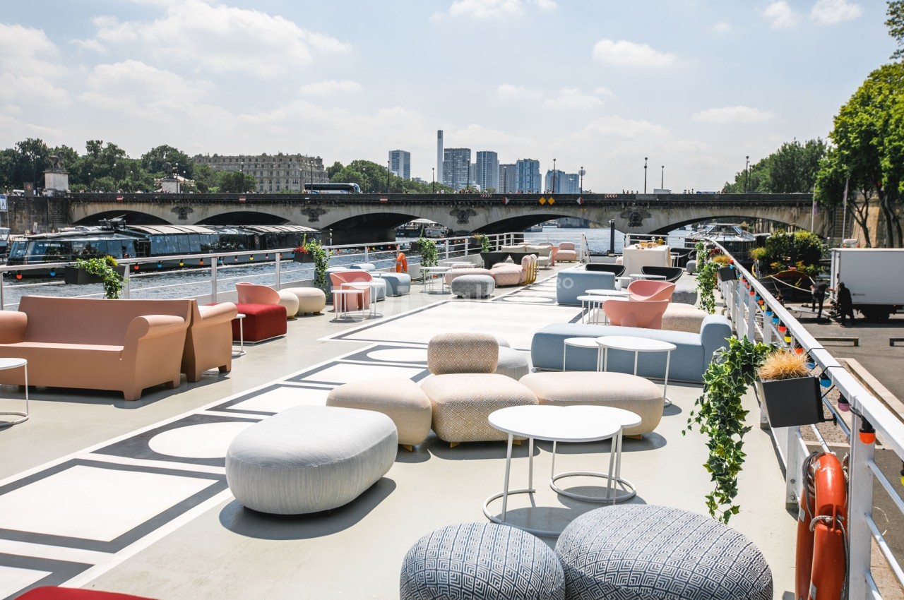 Terrasse aménagée avec des sièges et tables modernes, située au bord d'une rivière avec un pont en arrière-plan.