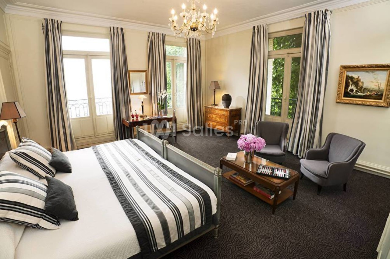 Chambre lumineuse avec lit double, fauteuils, table basse, commode, rideaux rayés et lustre au plafond.