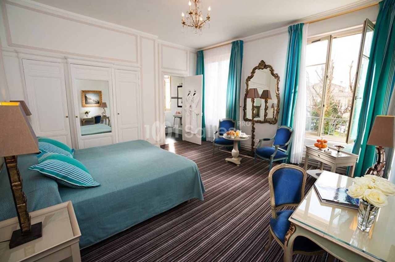Chambre lumineuse avec lit double, fauteuils bleus, bureau, miroir et grandes fenêtres donnant sur un extérieur arboré.
