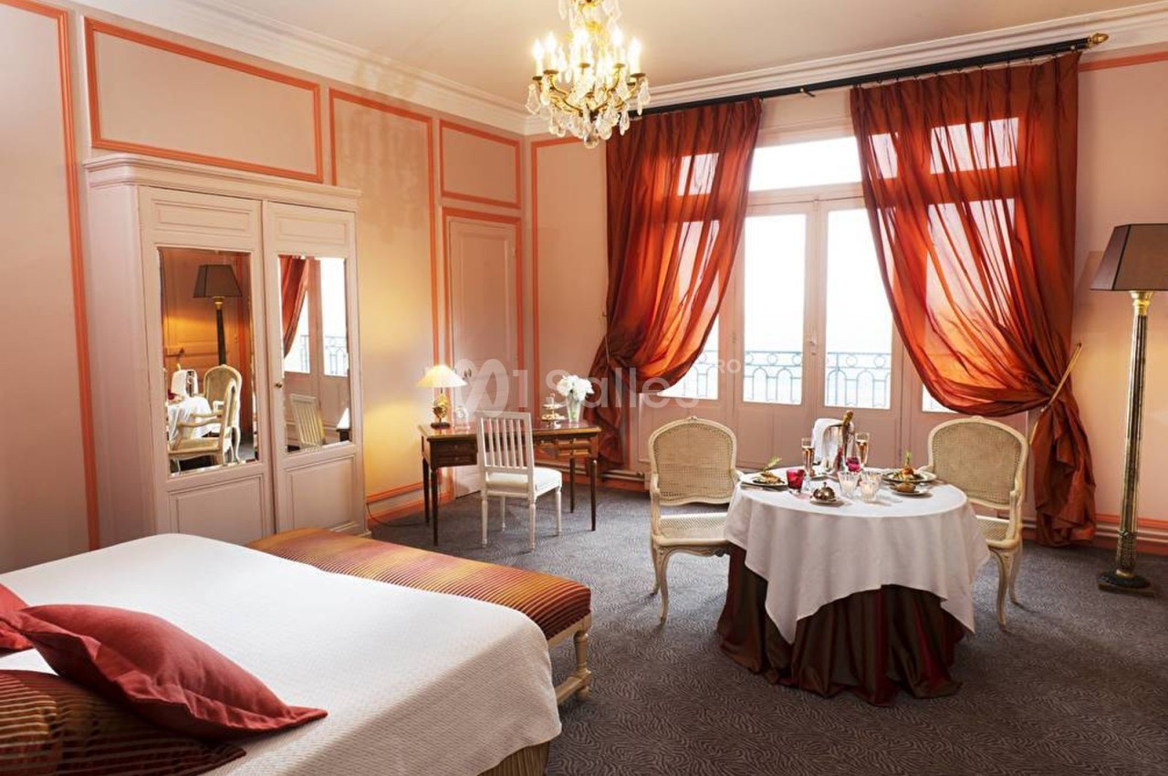 Chambre élégante avec lit double, table dressée pour un repas, grandes fenêtres et rideaux rouges.