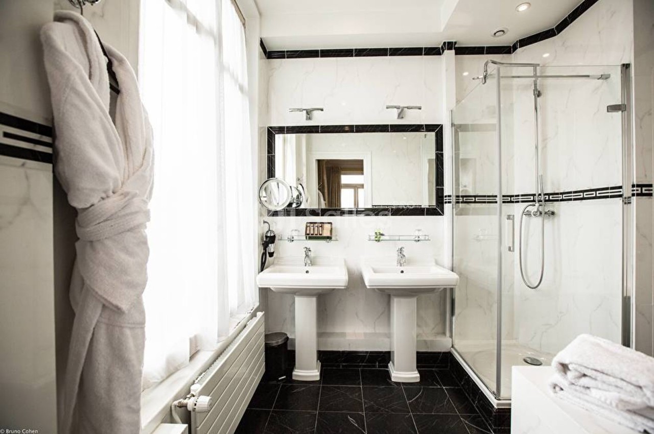 Salle de bain lumineuse avec deux lavabos, une douche vitrée, un peignoir suspendu et des murs en marbre blanc.
