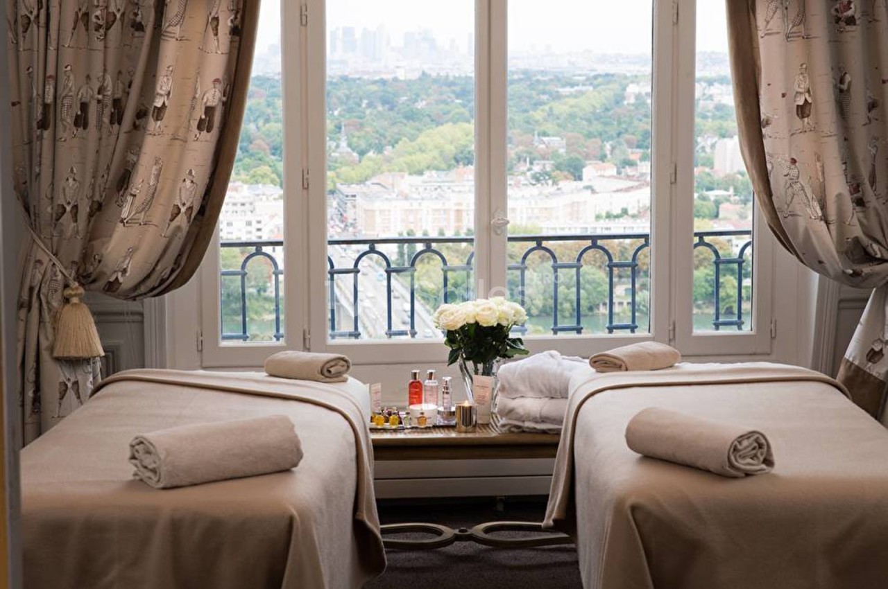 Deux tables de massage recouvertes de draps beiges, avec une vue sur un paysage urbain et verdoyant à travers une fenêtre.