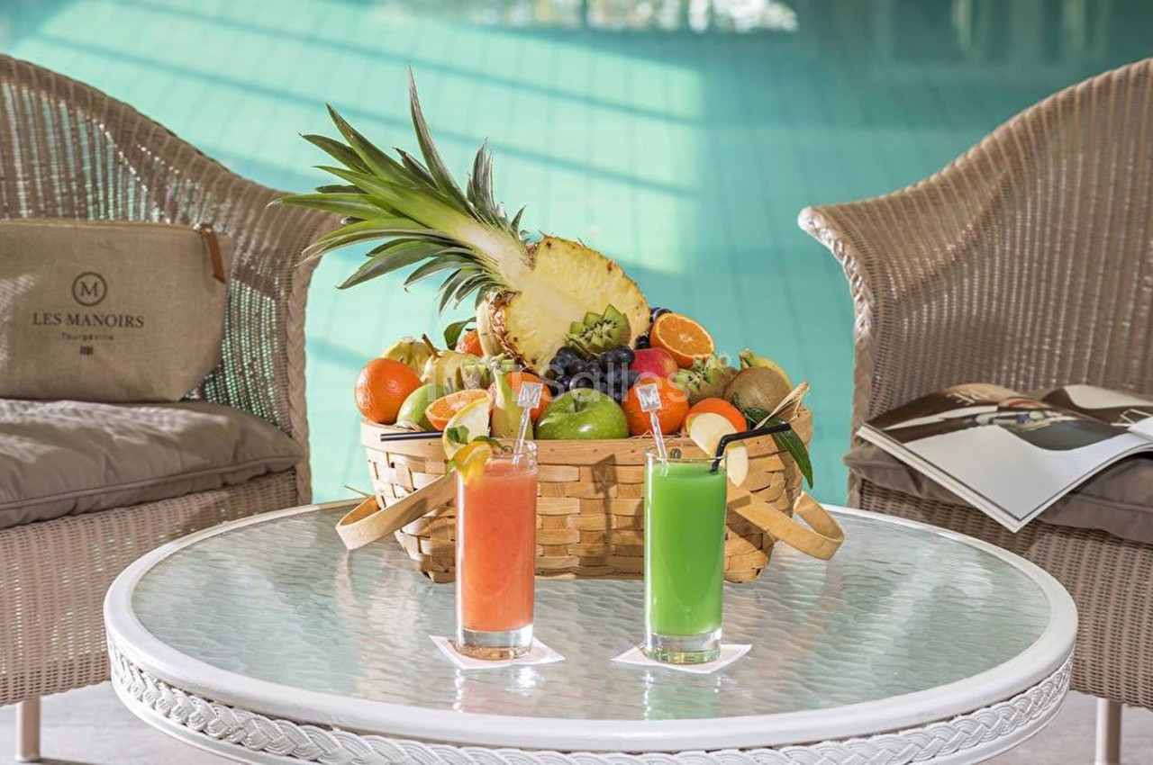 Panier de fruits frais sur une table en verre avec deux cocktails colorés, entouré de fauteuils en rotin.