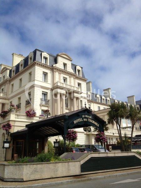 Façade d'un grand hôtel de style classique avec balcons fleuris, marquise en fer forgé et ciel partiellement nuageux.