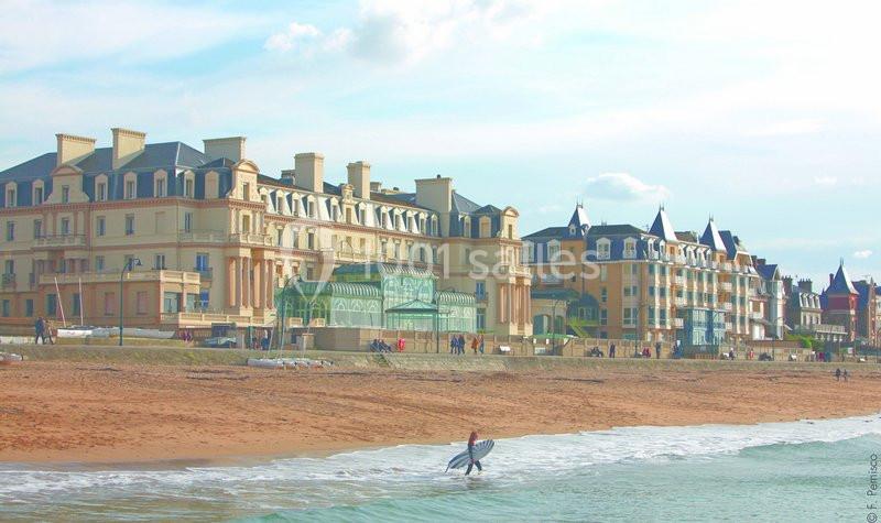 Un surfeur sort de l'eau sur une plage devant des bâtiments élégants de style balnéaire.
